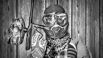 Ein detailreiches Schwarz-Weiß-Foto eines Paintballspielers, der eine Schutzmaske und ein gemustertes Halstuch trägt. Er hält sein Paintball-Markierer fest, und die Spiegelung in seiner Maske lässt Andeutungen von Reflektionen erkennen. Der Hintergrund besteht aus vertikalen Holzbrettern.